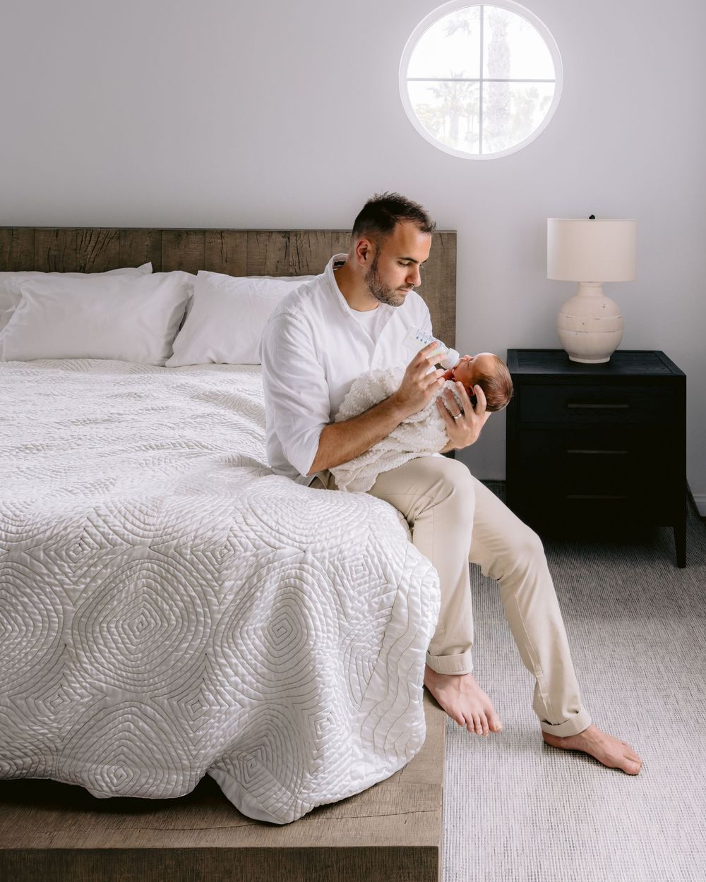 Person in light clothing sits on edge of white bed while looking at phone in minimalist bedroom.