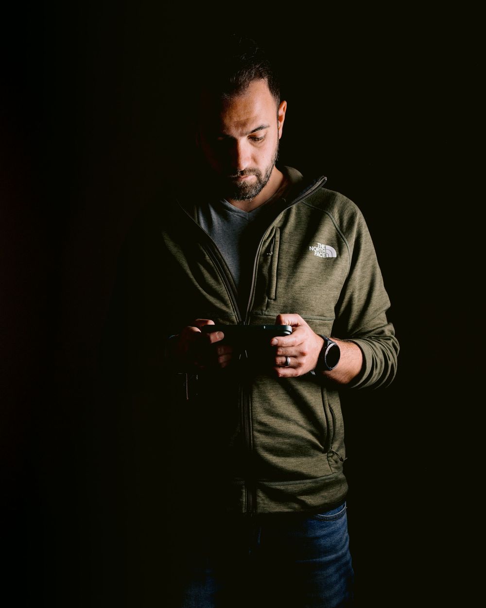 Silhouetted figure in dark hoodie standing against black background while looking at mobile device.