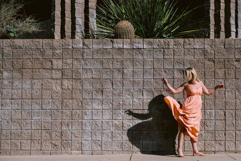 Woman in a coral dress walks along a stone wall with palm trees and greenery in a serene outdoor setting.