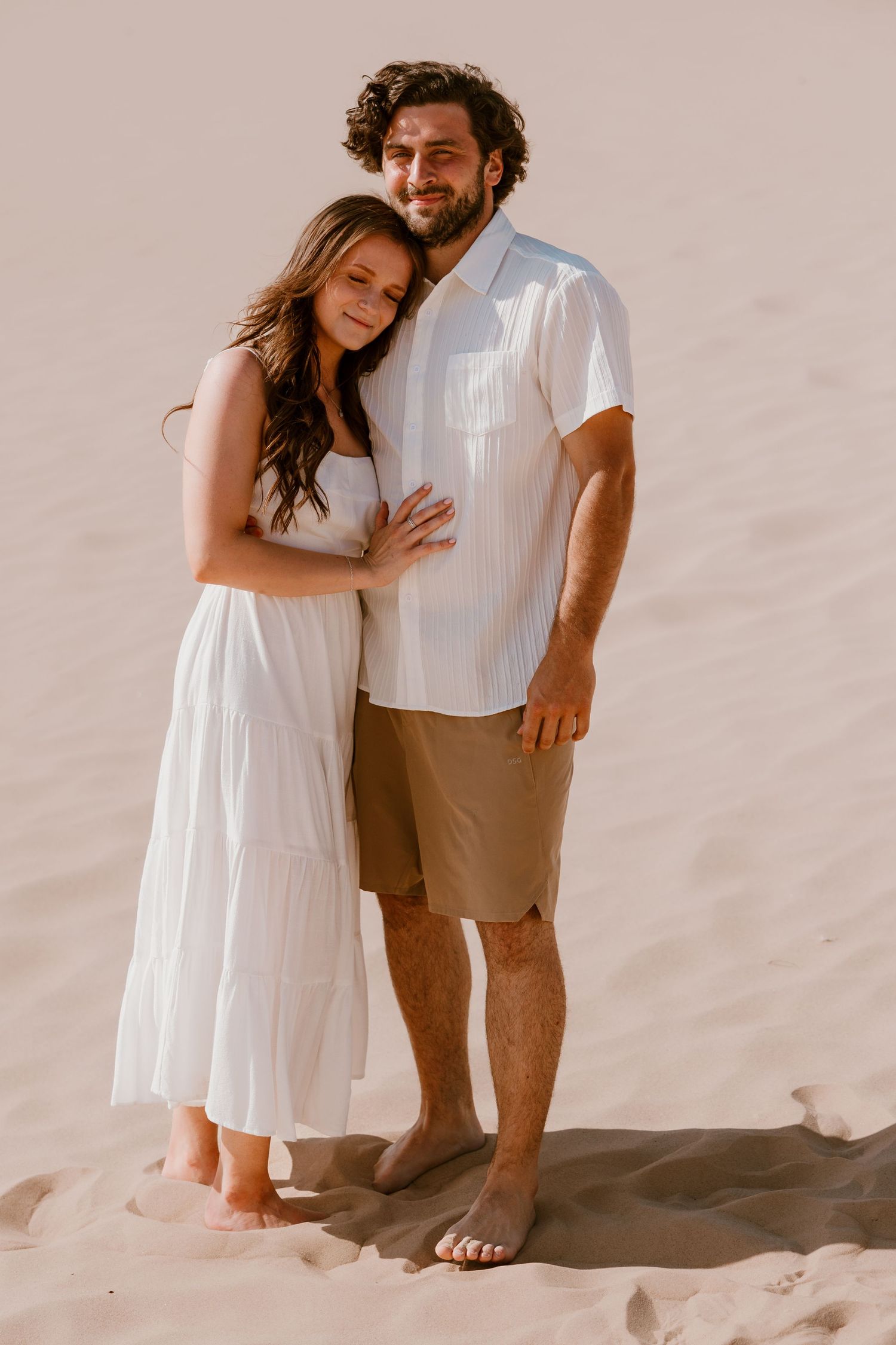 Couple in white outfits sharing intimate candid moments on a sandy beach in warm lighting.