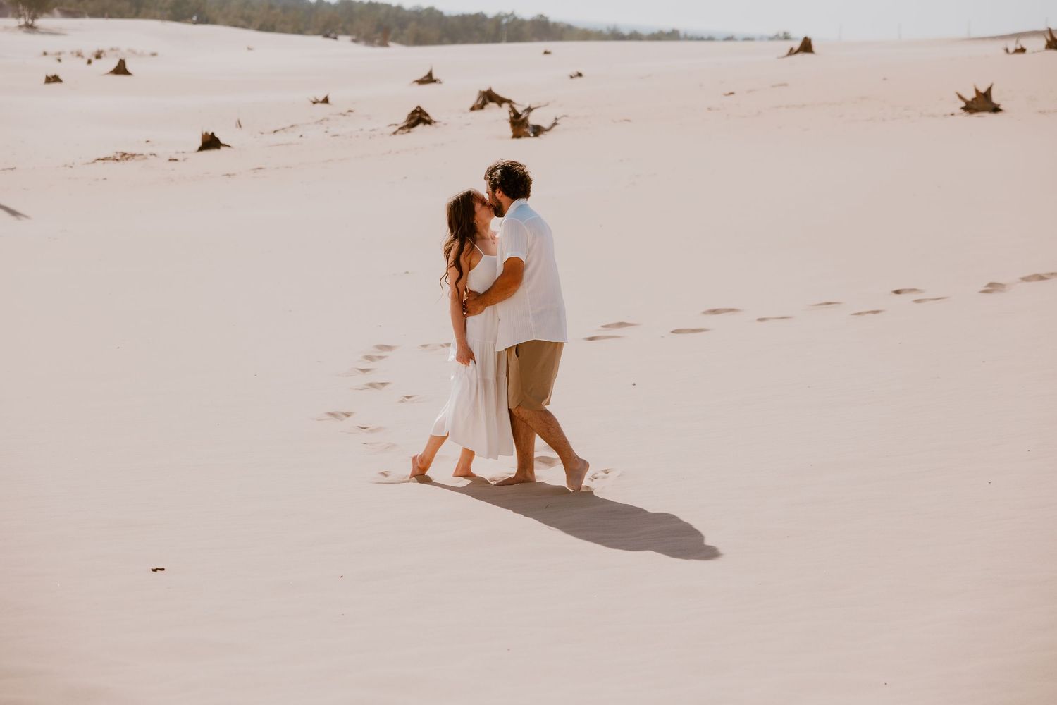 Two people walk hand in hand along a sandy beach shoreline in soft summer light creating romantic silhouettes.