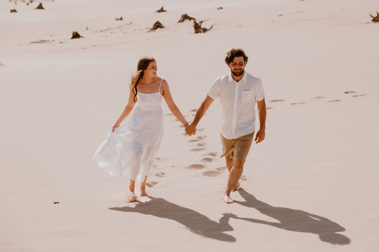 A couple walks hand in hand along a sandy beach, casting long shadows as the woman's white dress flows in the wind.