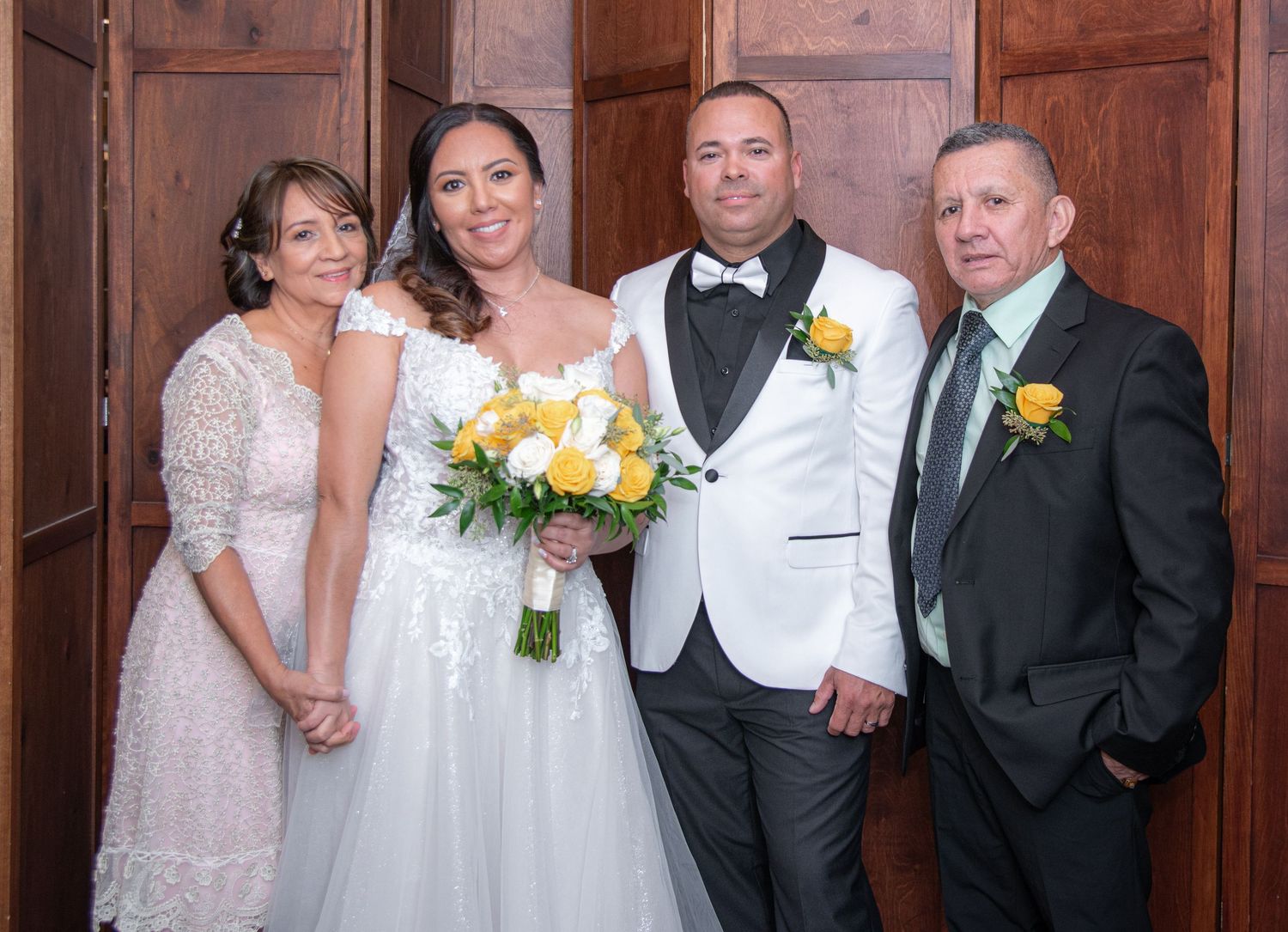 A wedding party in formal attire poses together against a wood-paneled wall at a celebration.