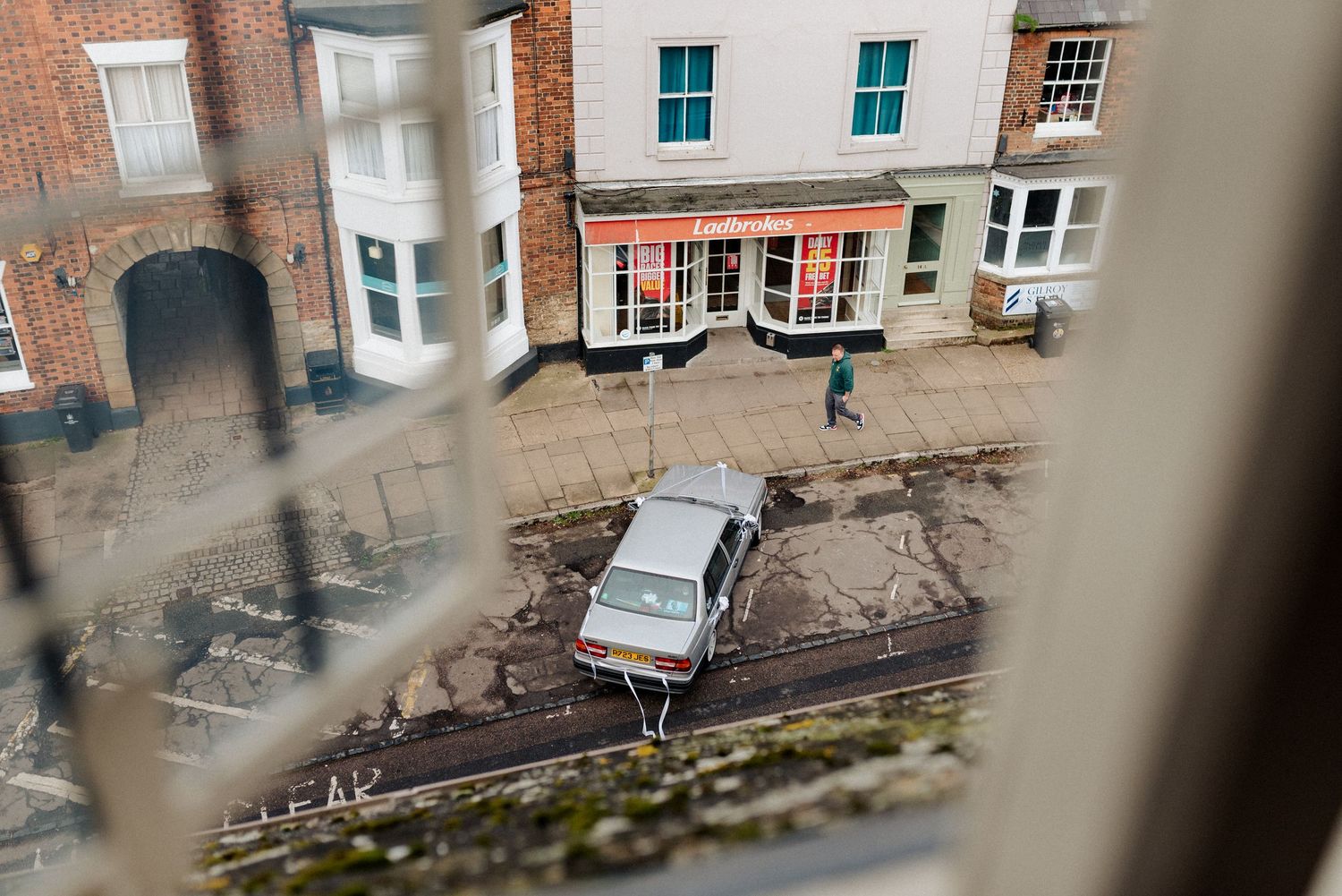 Bird's eye view through window bars of a vintage Volvo parked on a brick-lined street in front of historic storefronts.