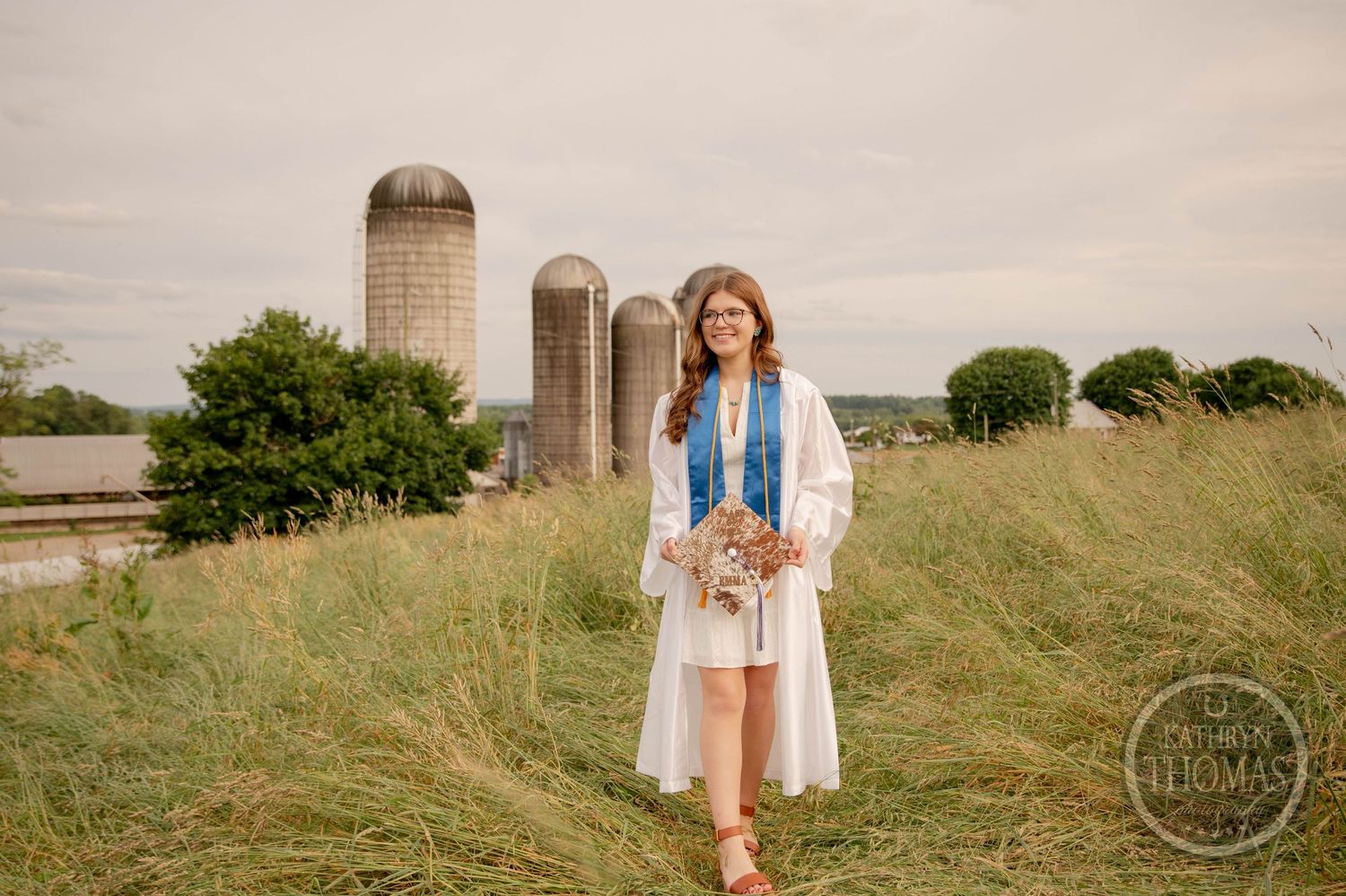 A person in a white dress and denim jacket poses in front of rustic farm silos on a grassy hill.