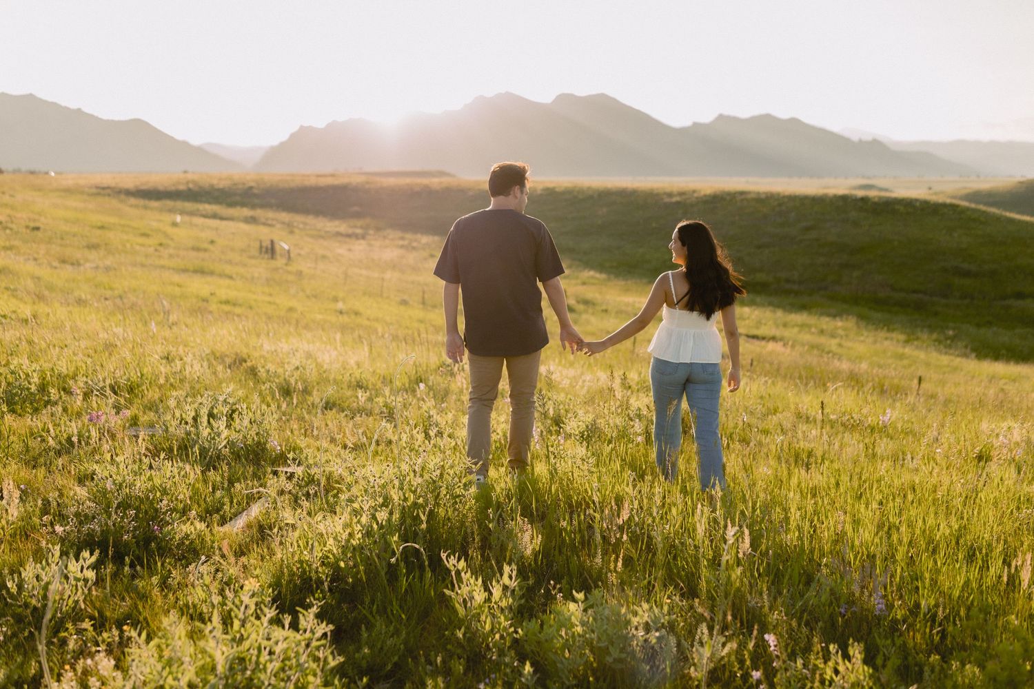 Two people walk hand-in-hand through a golden meadow at sunset with mountains in the distance.