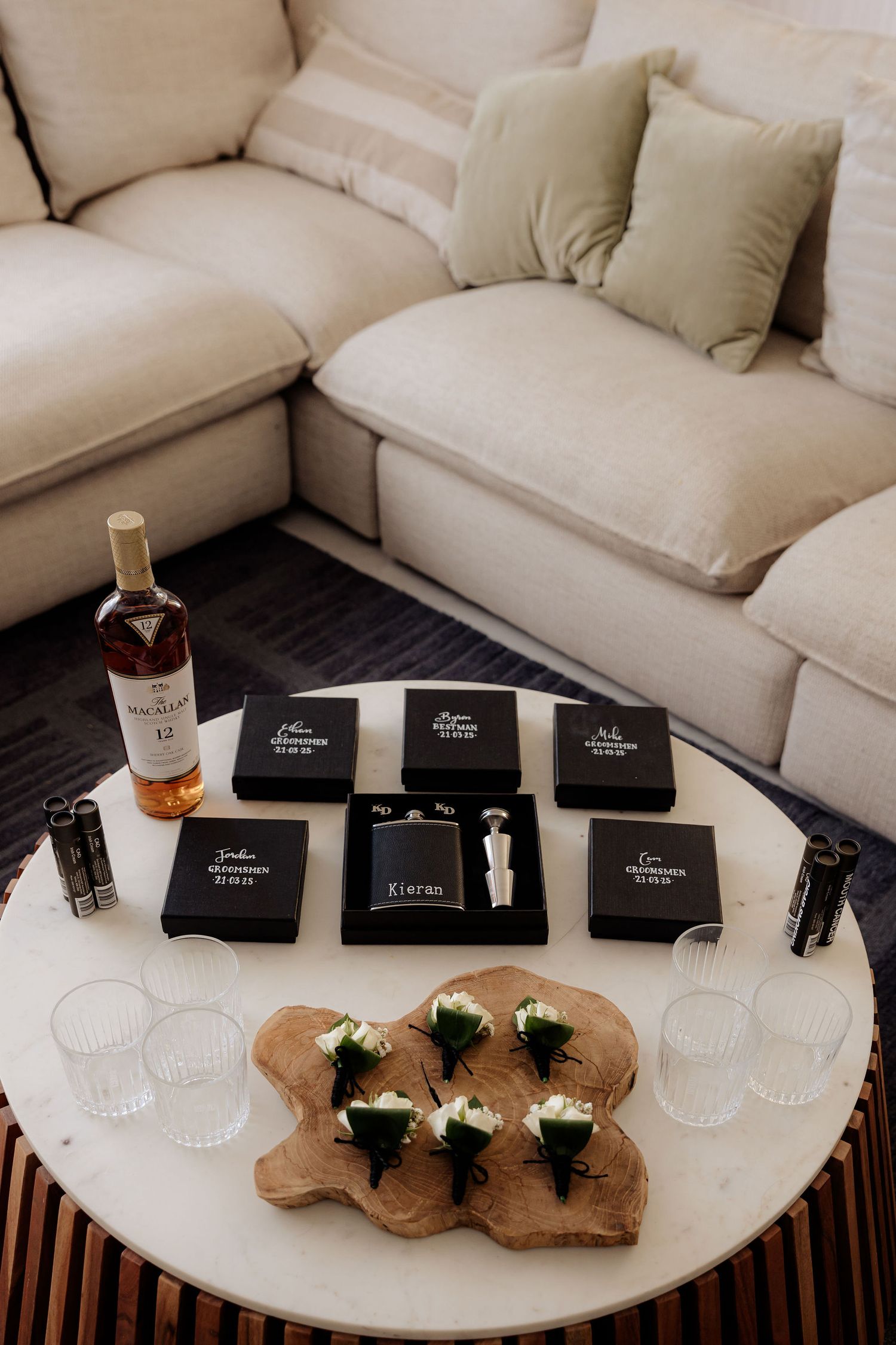 Wedding accessories including boutonnières, gift boxes, and a whiskey bottle arranged on a round table near a beige sectional sofa.