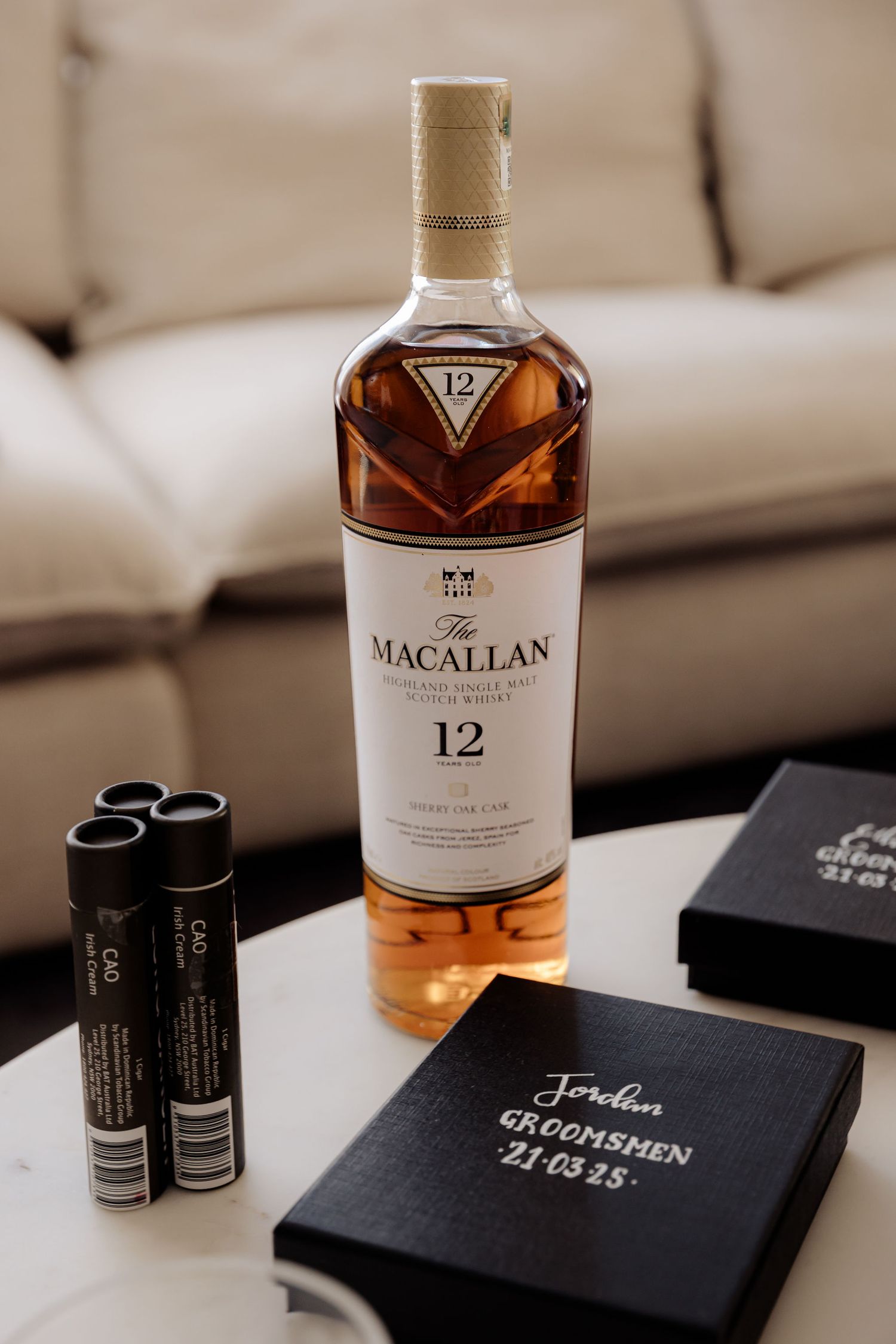 A bottle of Macallan 12-year scotch whisky sits on a table with black notebooks and two black tubes in a cozy setting.