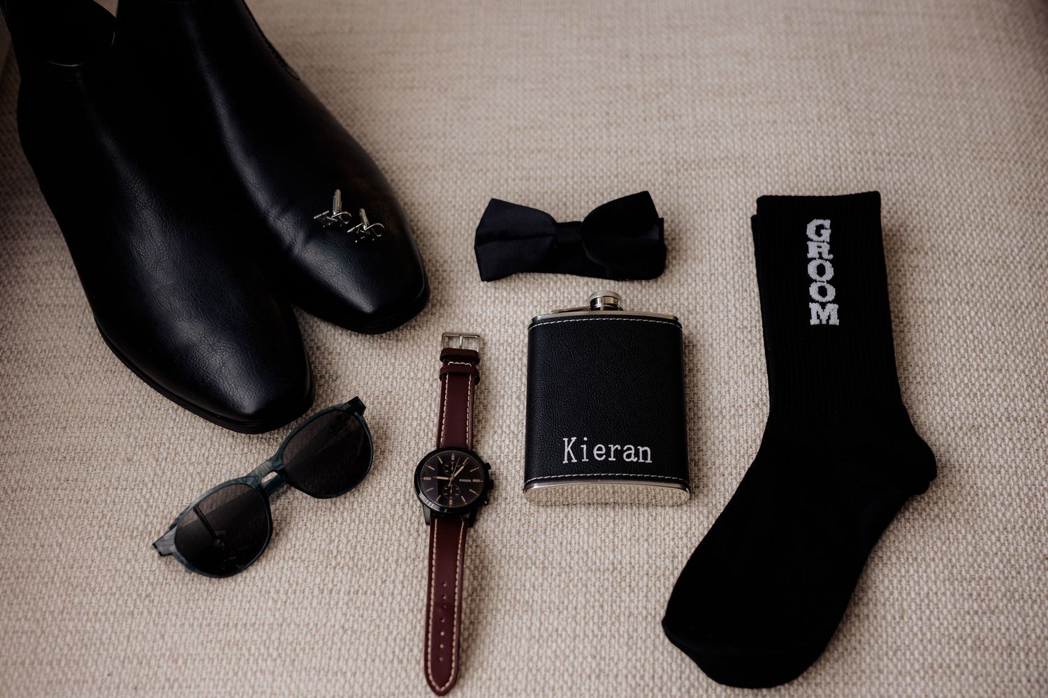 Groom's wedding accessories including black shoes, bowtie, sunglasses, leather watch, flask, and black socks on beige fabric.