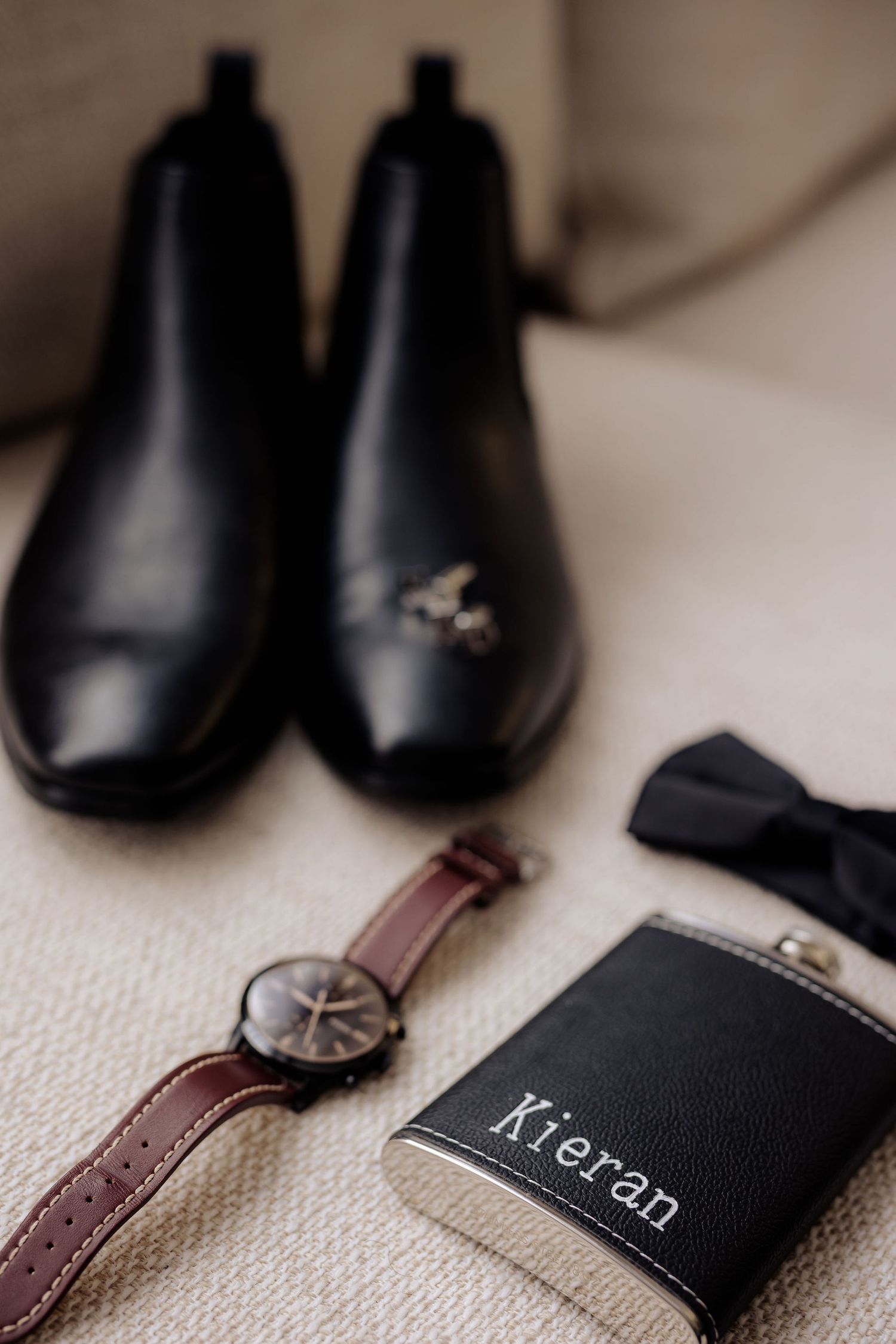 A groom's accessories including black leather shoes, brown leather watch and personalized black hip flask on beige fabric.