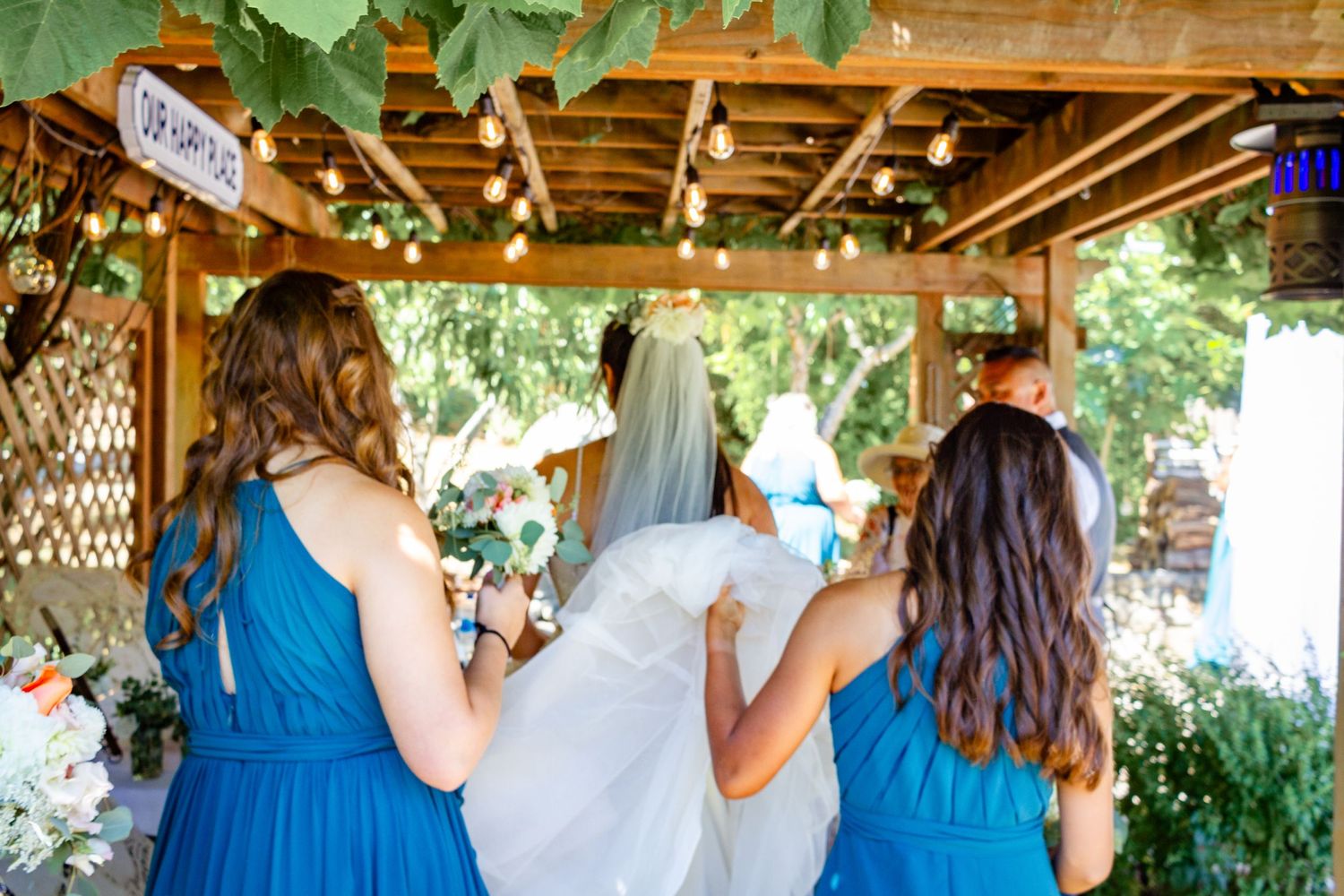 Bridesmaids in blue dresses gather under a wooden pergola at an outdoor wedding venue.