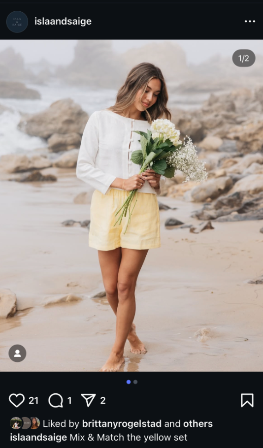 Woman in white top and yellow linen shorts holding white flowers on a rocky beach at sunset.