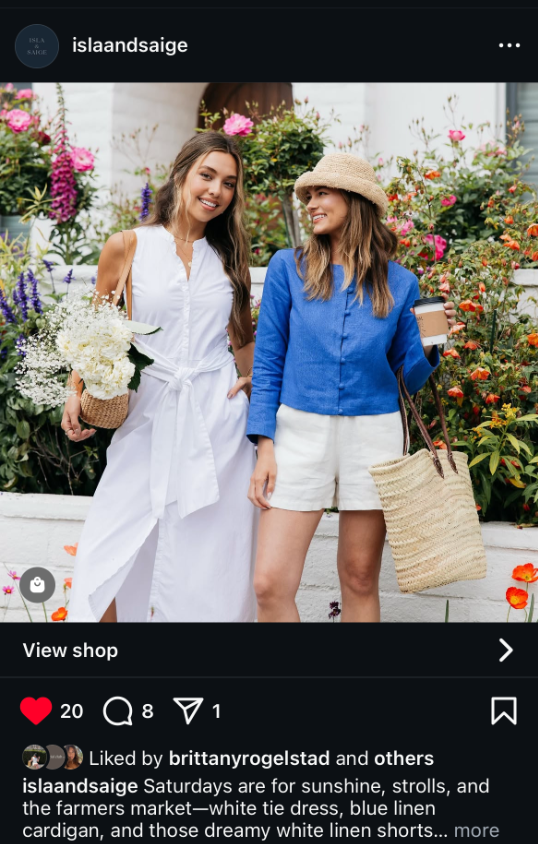 Two people in casual summer outfits with straw bags pose against a colorful garden backdrop of pink and white flowers.