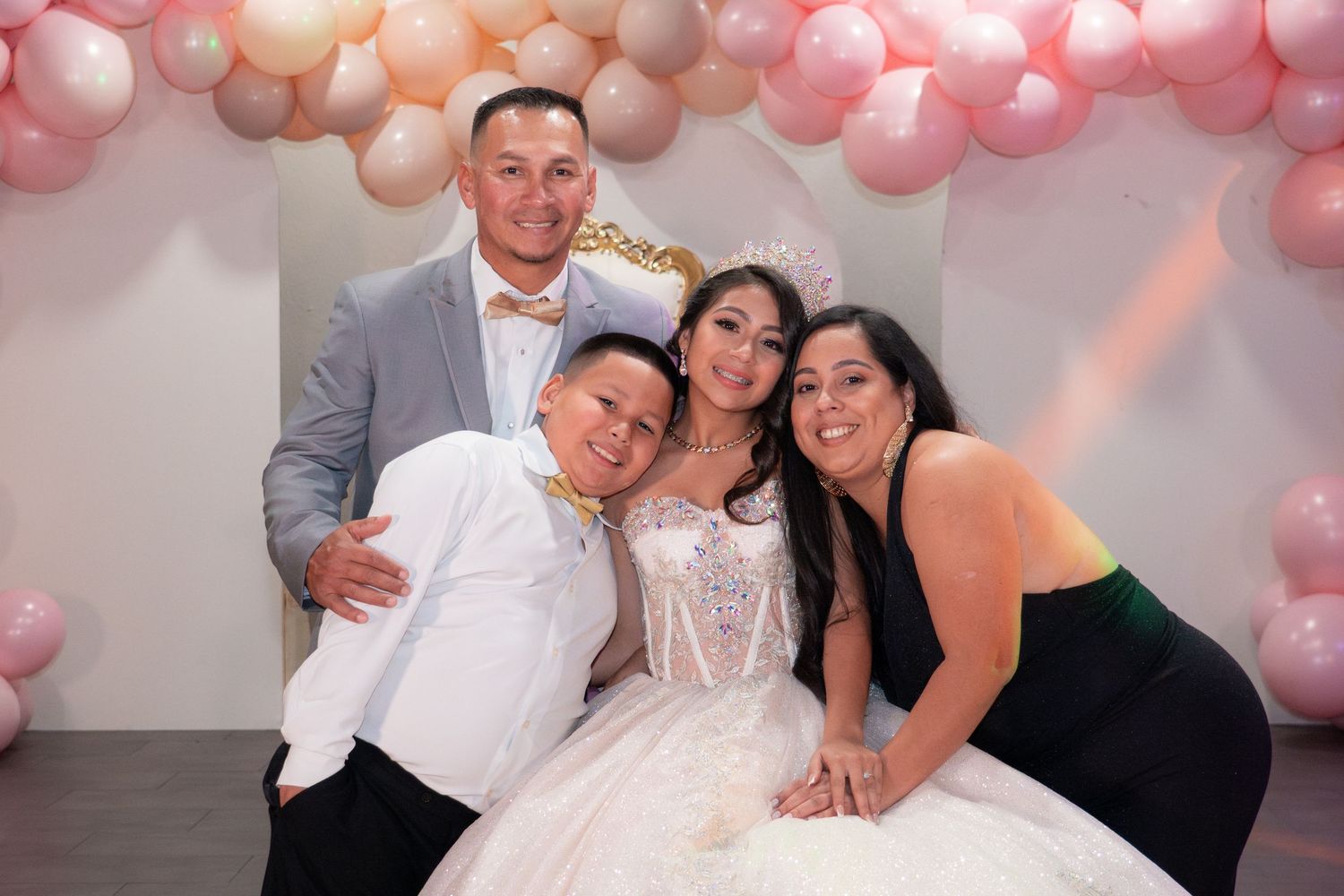 Family of four poses together at a quinceañera celebration with pink balloon arch and white gown.