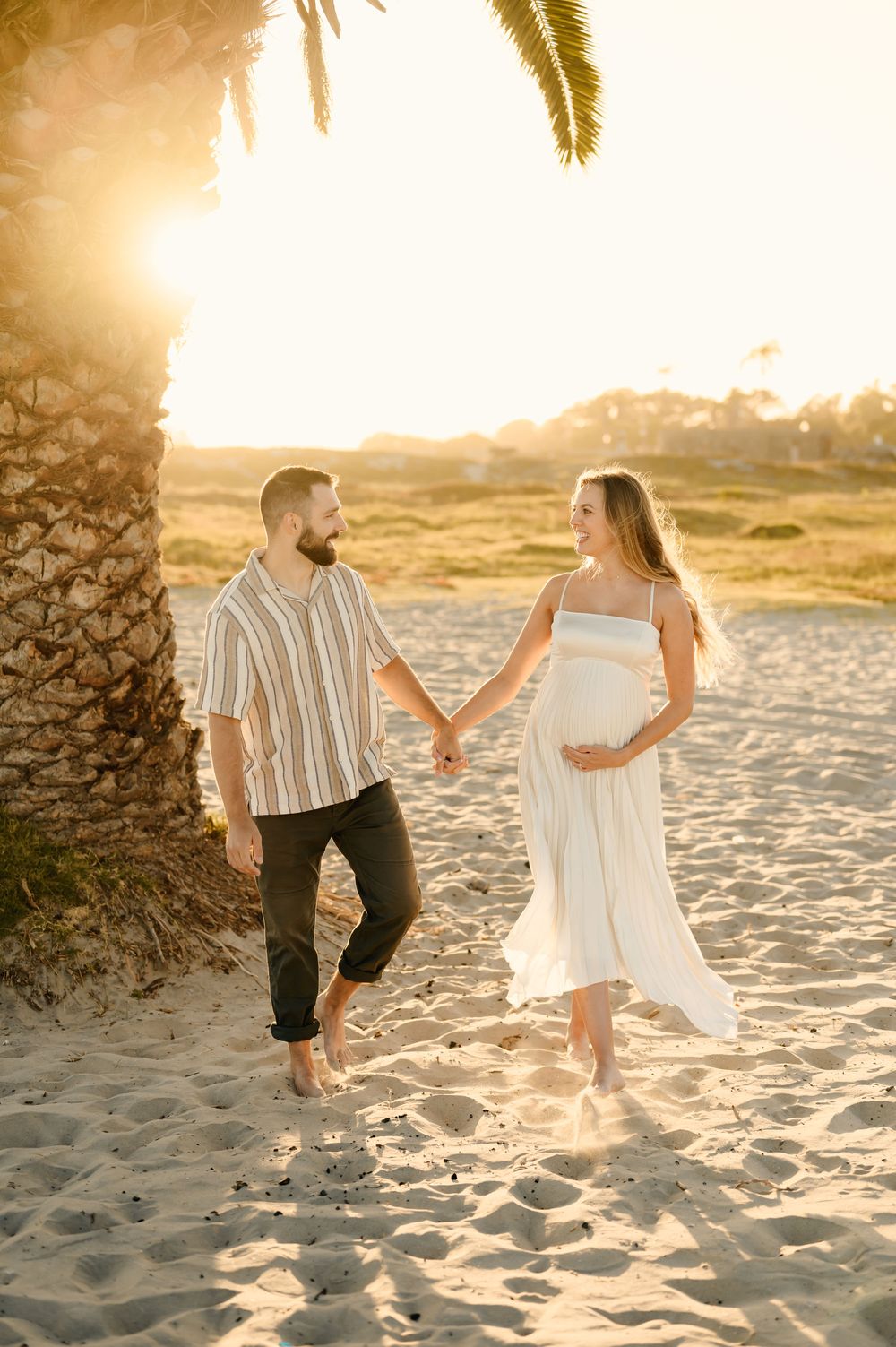 Couple holding hands while walking in sunshine under palm tree during their maternity photoshoot in Ventura