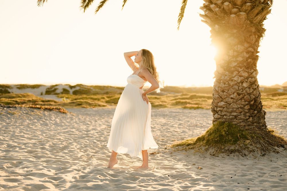 woman poses in sunshine in white dress highlighting her silhouette during her maternity photoshoot by Ventura photographer