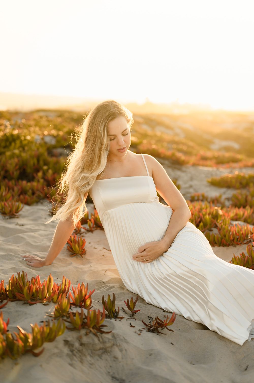 woman lays on succulent covered sand dunes on coronado beach holding her belly during her maternity photoshoot by Ventura photographer