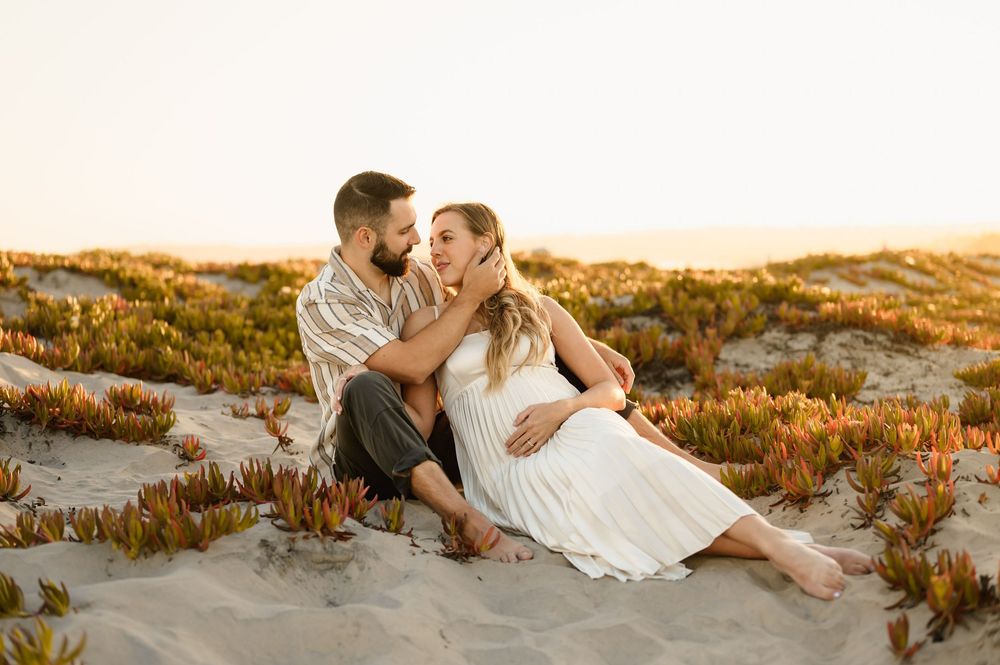 Couple lays together in succulent covered sand dunes on coronado beach during their maternity photoshoot by Ventura photographer