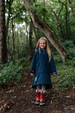 A person in a navy blue raincoat and red boots stands on a forest trail surrounded by greenery.