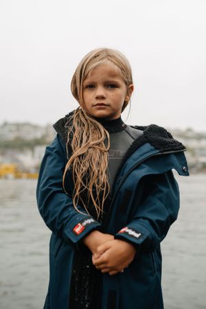 A person in a navy blue coat stands against an overcast harbor backdrop.