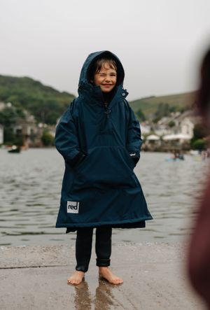 A person wearing a blue hooded raincoat stands barefoot on a wet shoreline.