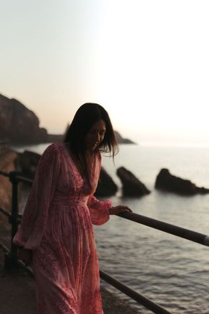 A person in a flowing pink dress stands by the ocean at sunset among rocky coastal cliffs.