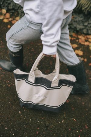 Close-up of legs wearing boots next to a white and navy canvas tote bag on wet ground.