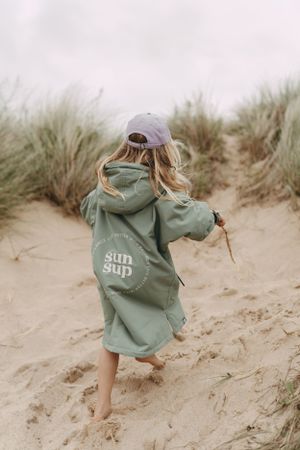 A child in a green hoodie running barefoot through sand dunes on a cloudy day.  Branding photography, Devon