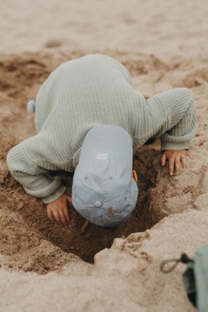 A child digging in the sand wearing a light-colored knit outfit and cap.