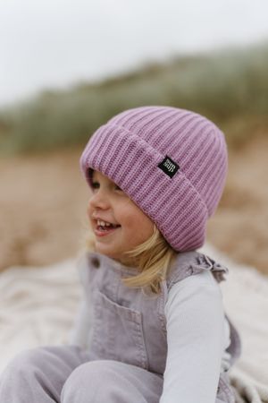 Child wearing a pink knitted beanie hat smiles while sitting outdoors on a cloudy day.