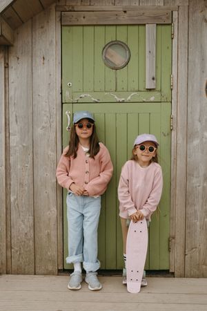 Two children in pink sweaters and sunglasses stand against a green wooden door holding a skateboard.