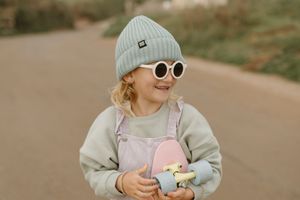 Young child in white sunglasses and gray beanie smiles while wearing overalls and holding a pink toy.