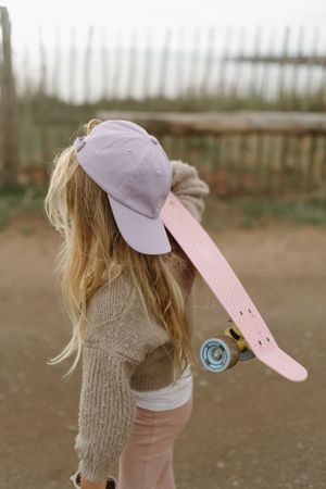 A skateboarder wearing a lavender cap and beige sweater carries a pink skateboard while standing outdoors.