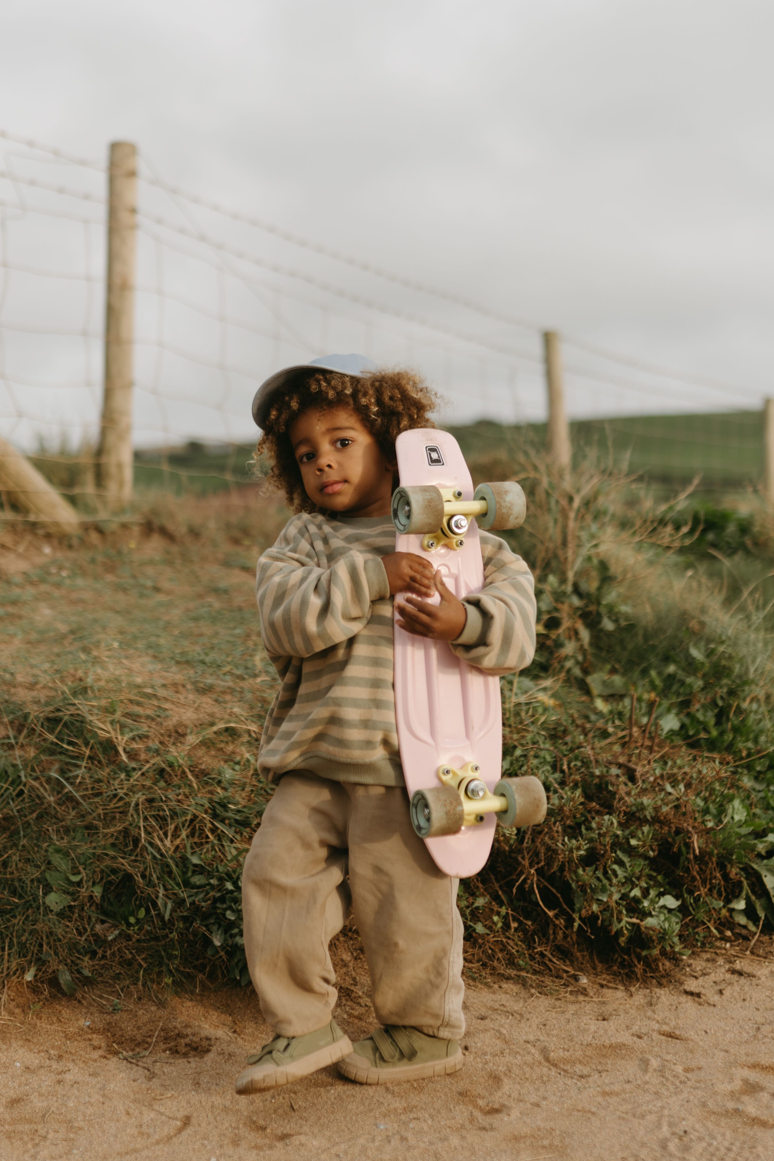 Young person in oversized clothing holding a pink skateboard beside a wire fence on a dirt path.