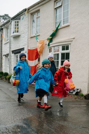 Children in bright raincoats running down a wet street past white houses with festive decorations.