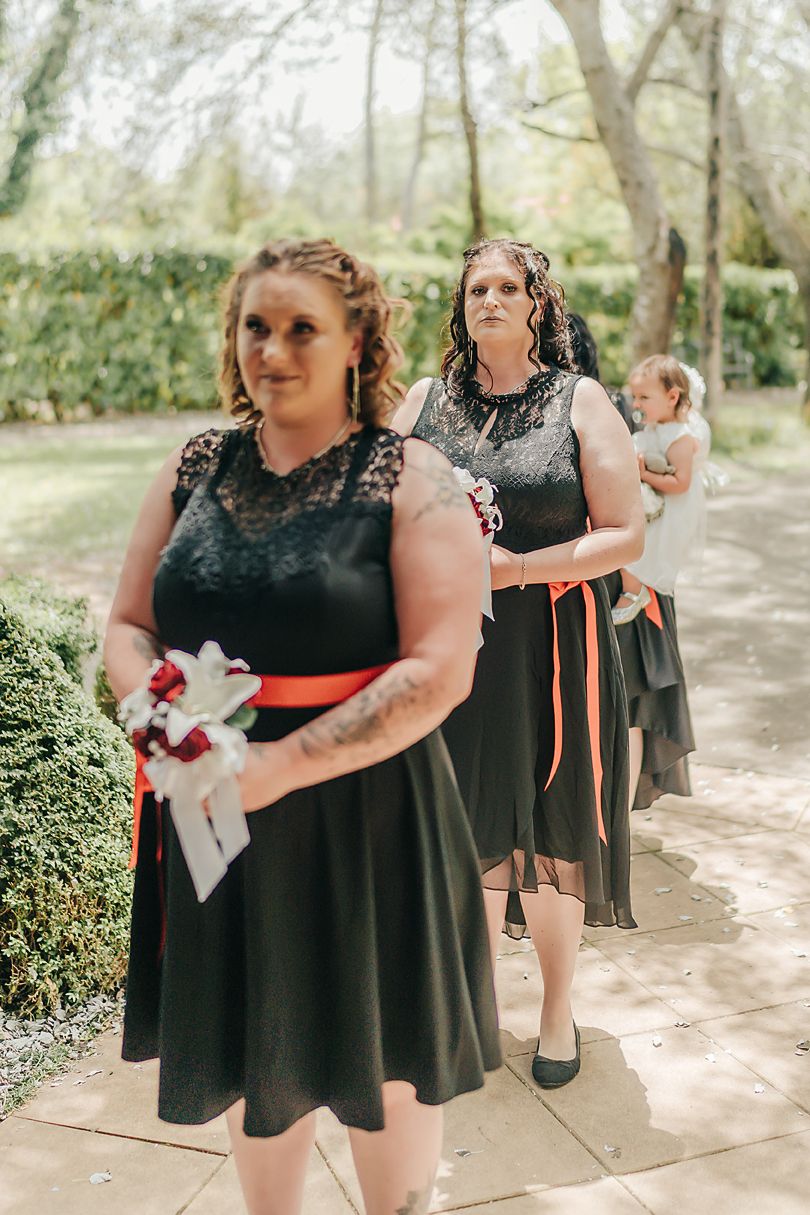 Bridesmaids in matching black knee-length dresses with orange sashes walk in line along garden path during outdoor wedding.