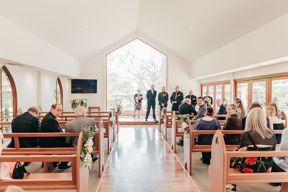 Guests seated in wooden pews inside a modern chapel with large windows and natural light streaming through.