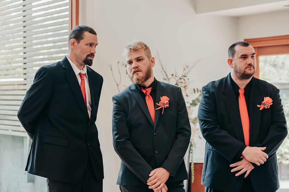 Three groomsmen in black suits with coral orange ties stand together at a wedding ceremony.