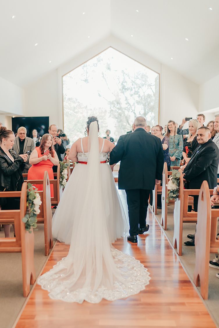 A bride in a flowing white gown walks down the church aisle with wooden pews and large windows letting in natural light.