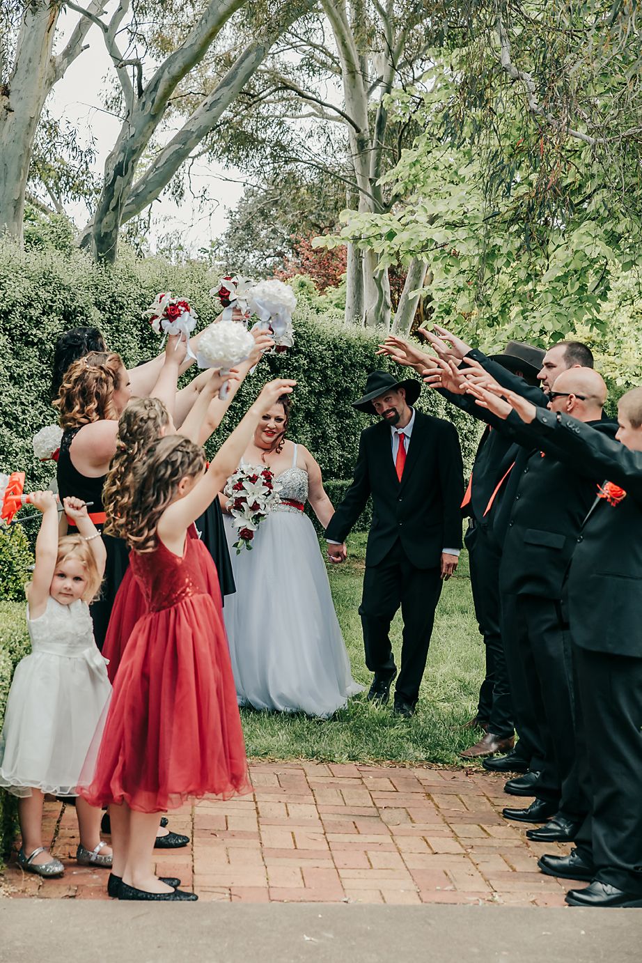 A wedding party celebrates joyfully on a brick path amidst lush garden greenery.