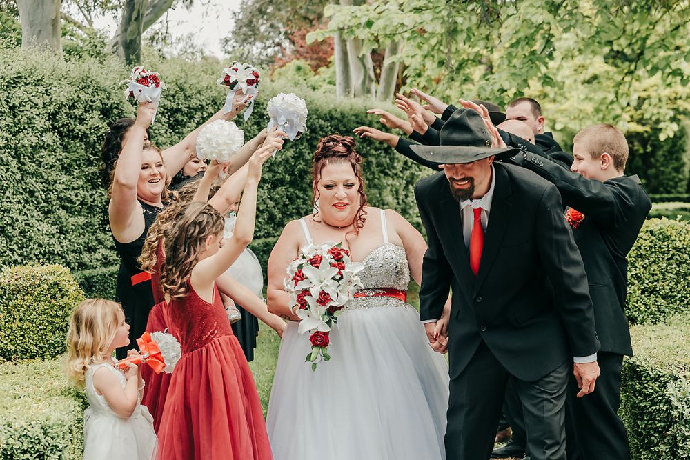 A wedding party celebrating outdoors in red bridesmaid dresses and black suits against a lush green garden backdrop.