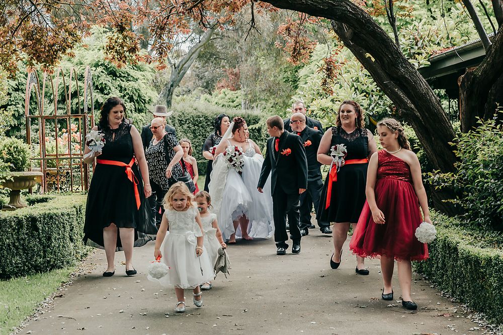 Wedding party walking down garden path with bridesmaids in red and black dresses outdoors.