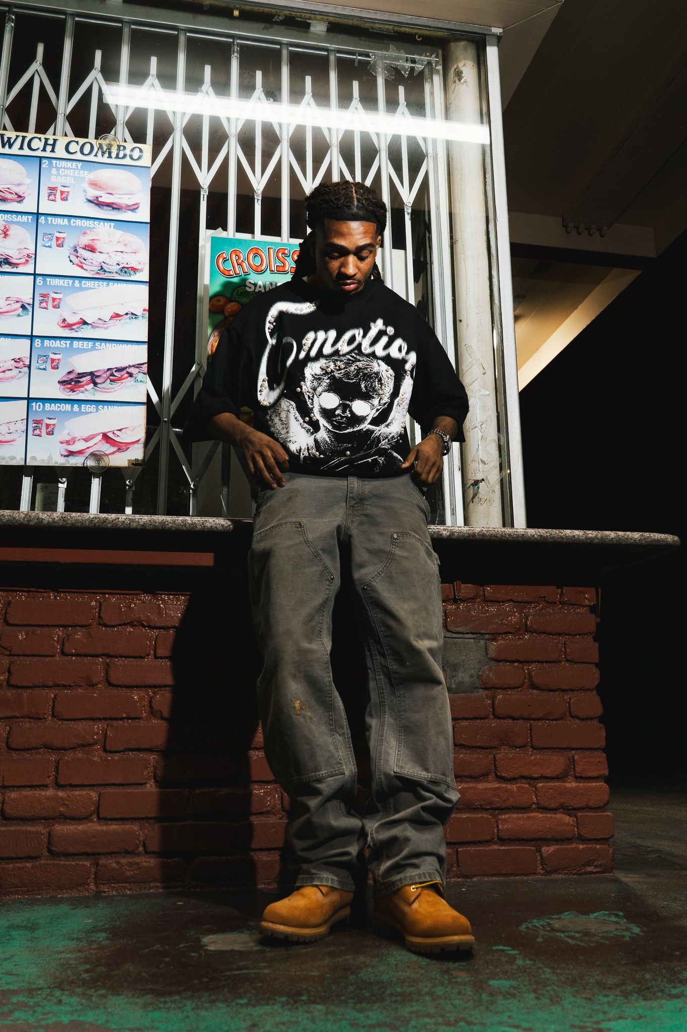 Person sitting on floor wearing baggy jeans and Timberland boots against a brick wall with illuminated signs above.