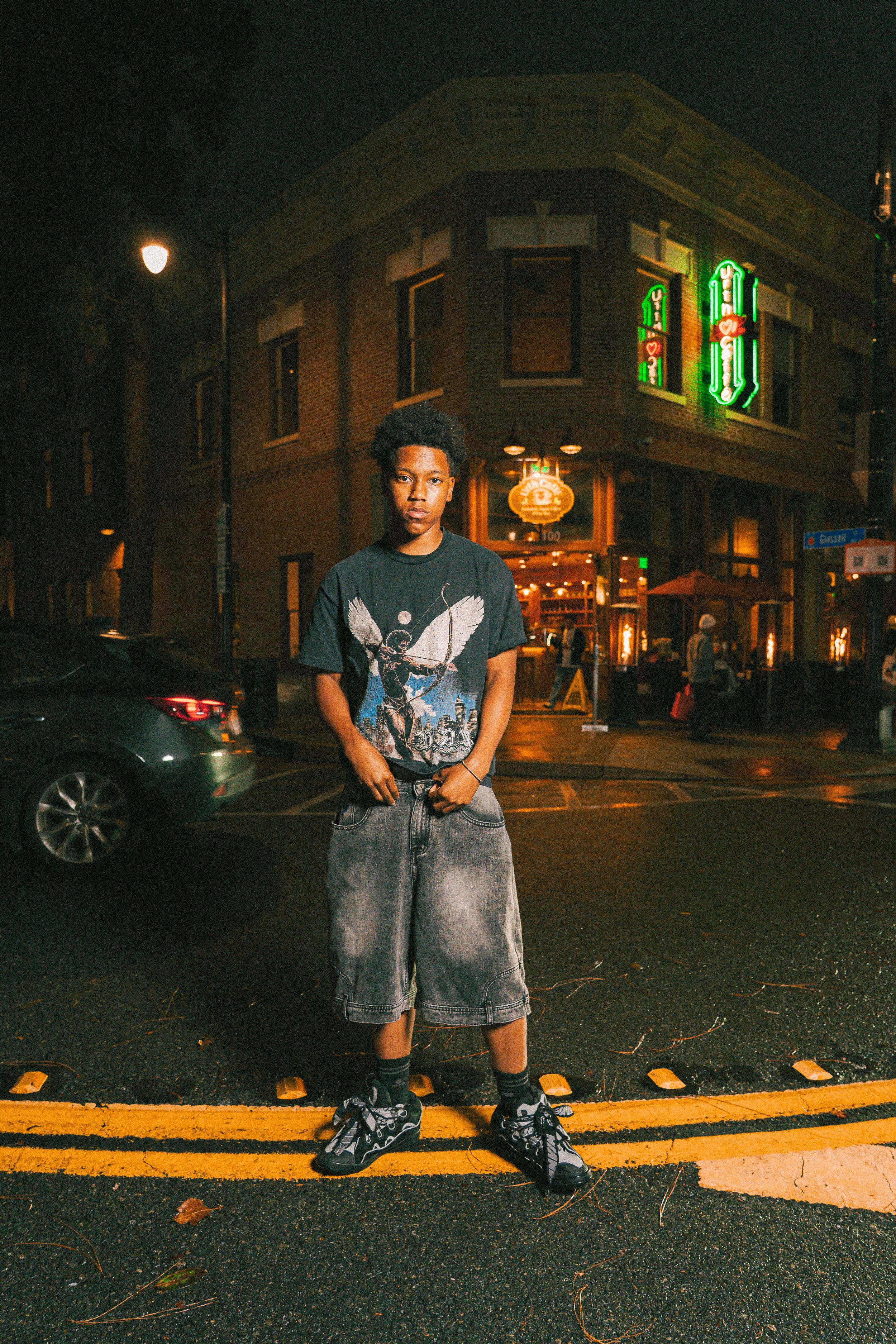 A person in a blue t-shirt poses with a scooter on a dark city street at night with orange lighting.