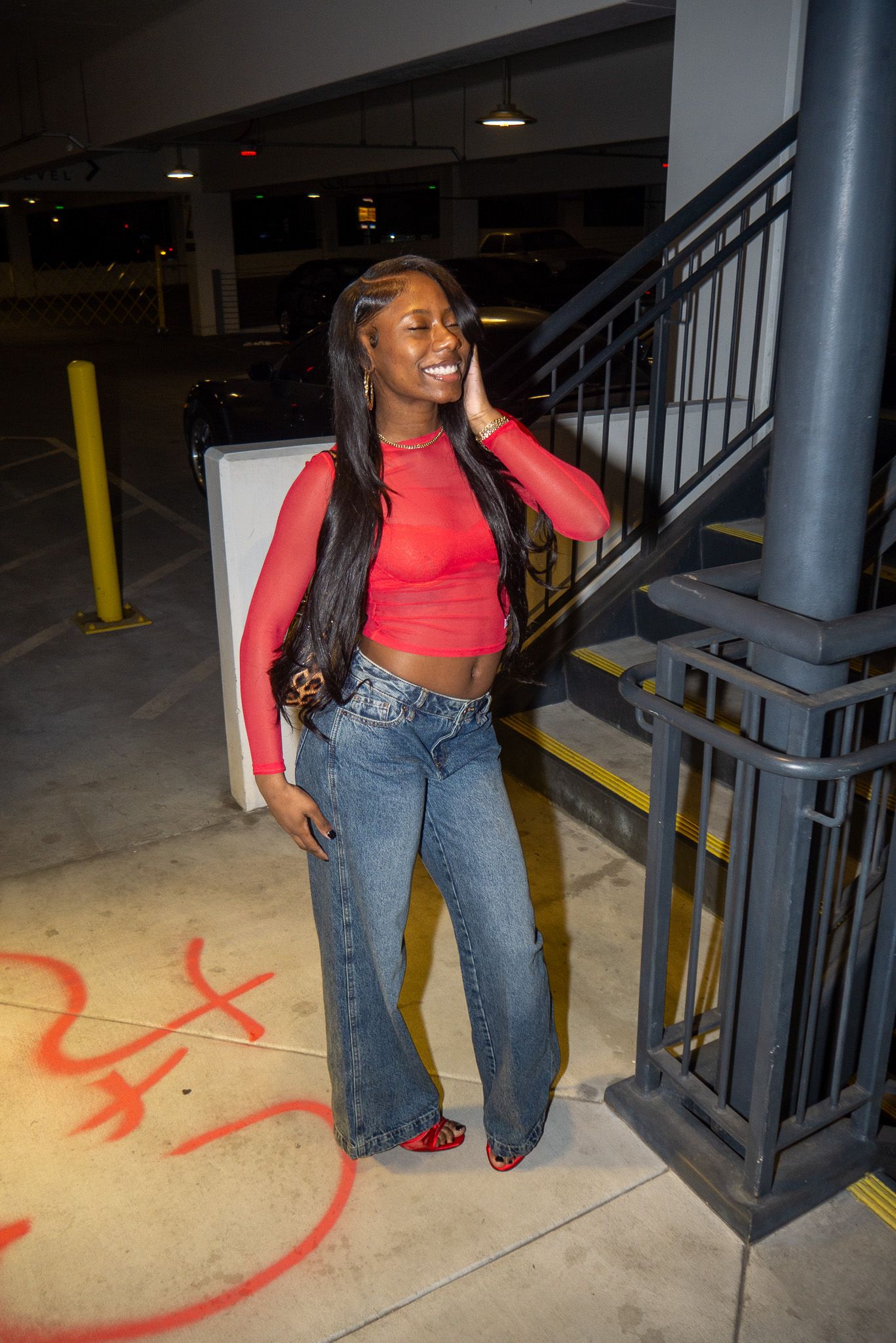 Person in red crop top and jeans poses playfully near concrete stairs in a parking structure.