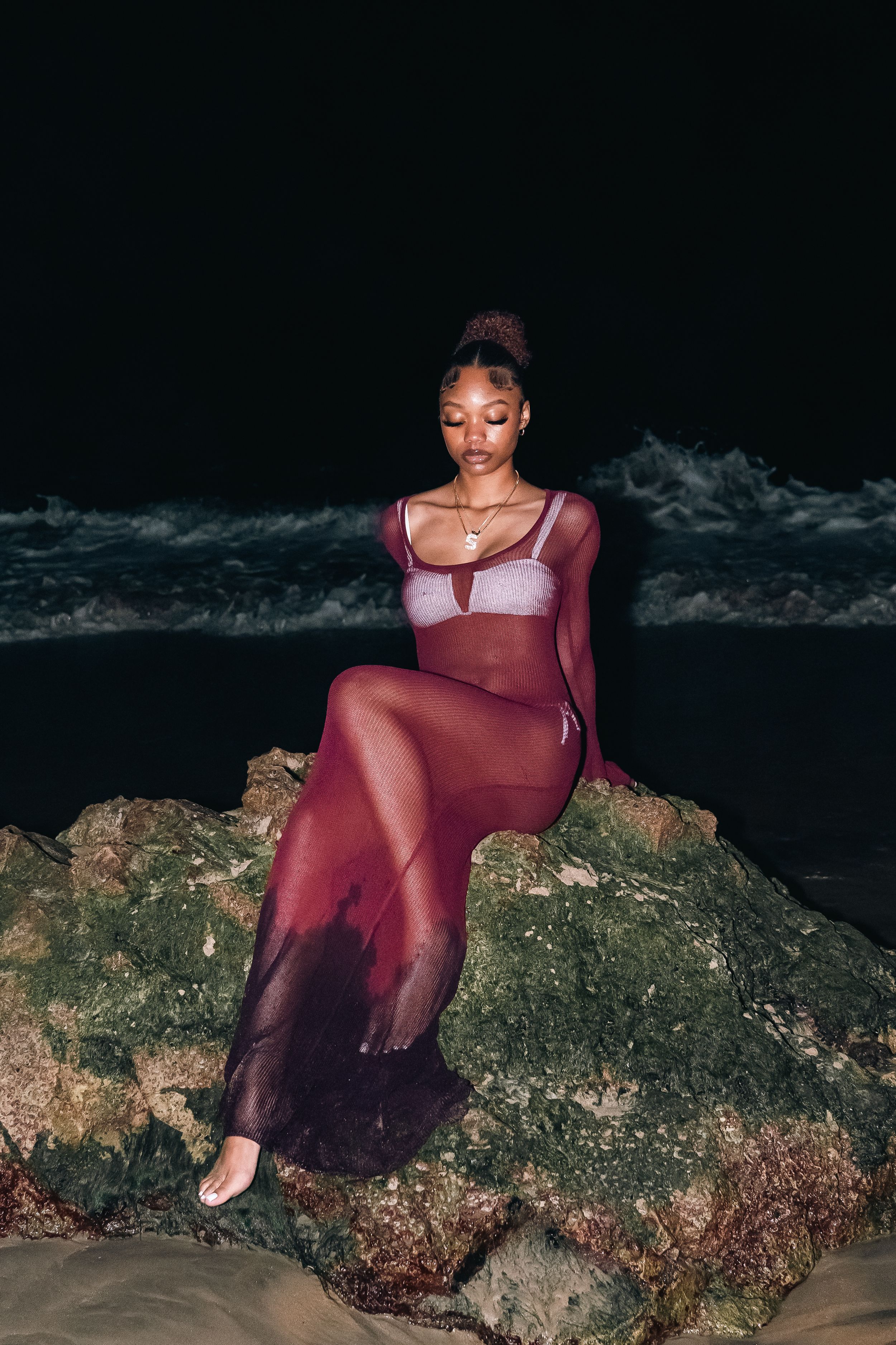 Person in burgundy sheer dress poses on rocky beach at night.