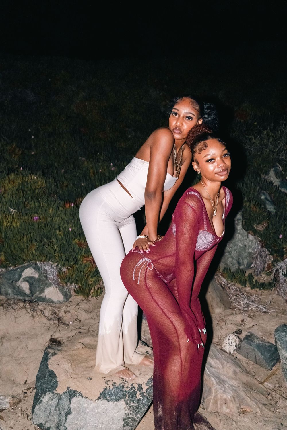 Two friends pose together on rocks, wearing white and burgundy evening outfits.