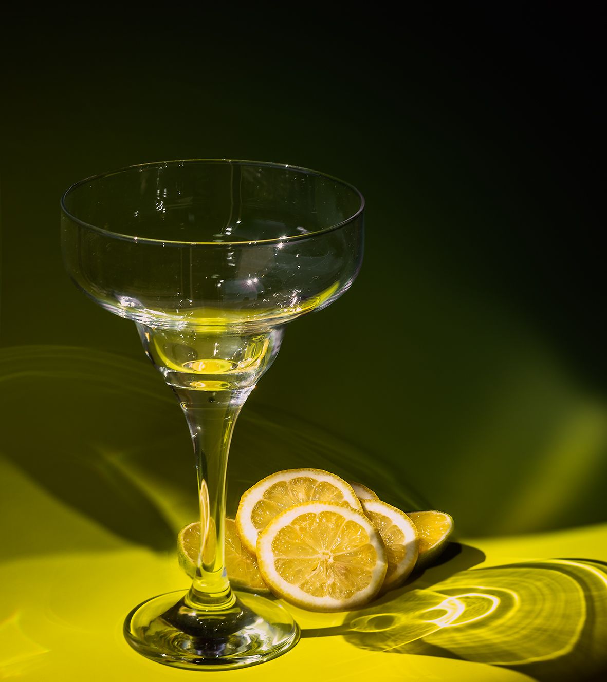 Empty cocktail glass with sliced lemons on yellow surface against dark background with dramatic lighting.