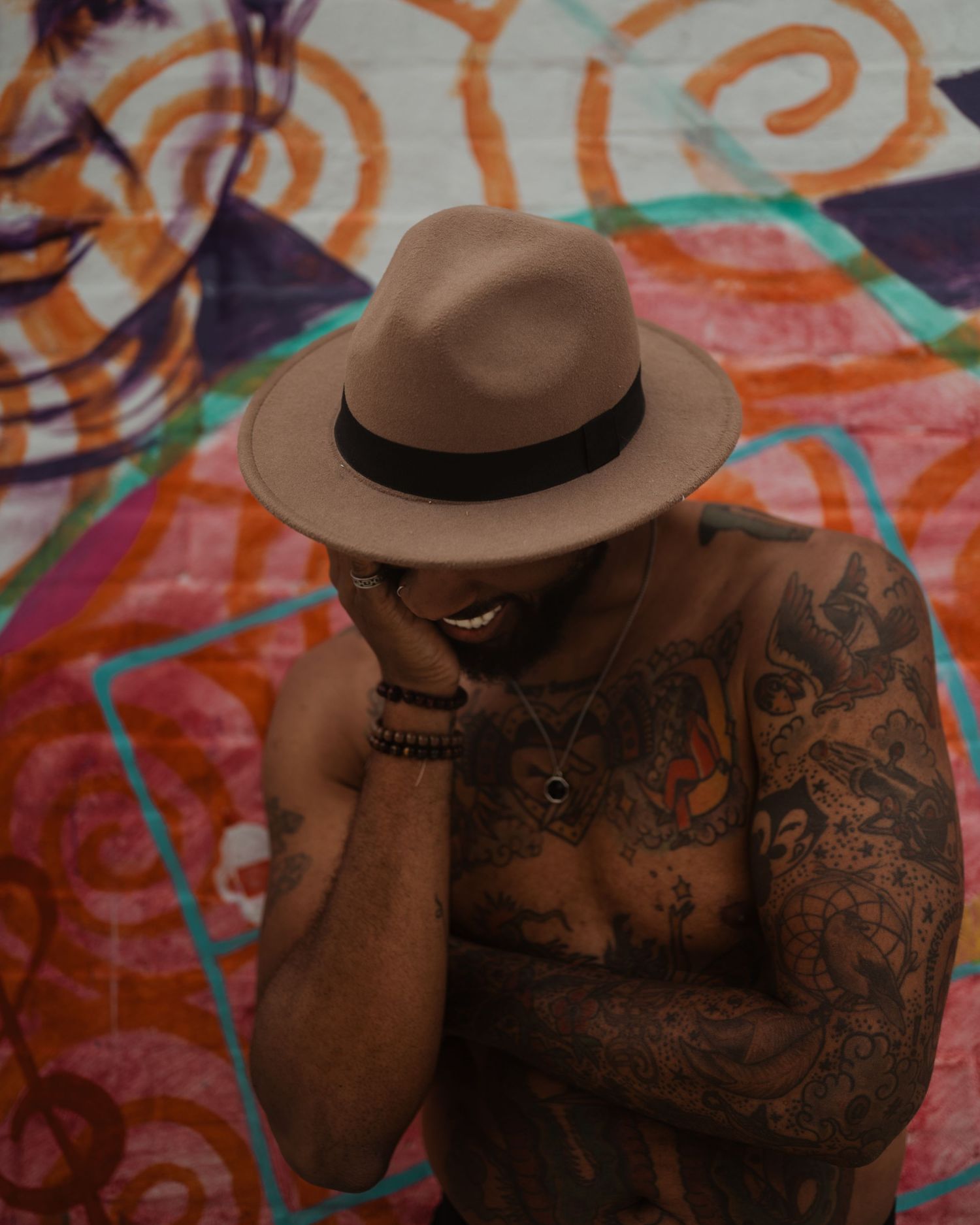 Person wearing brown fedora hat and showing detailed tattoo sleeve poses against colorful spiral wall art background.