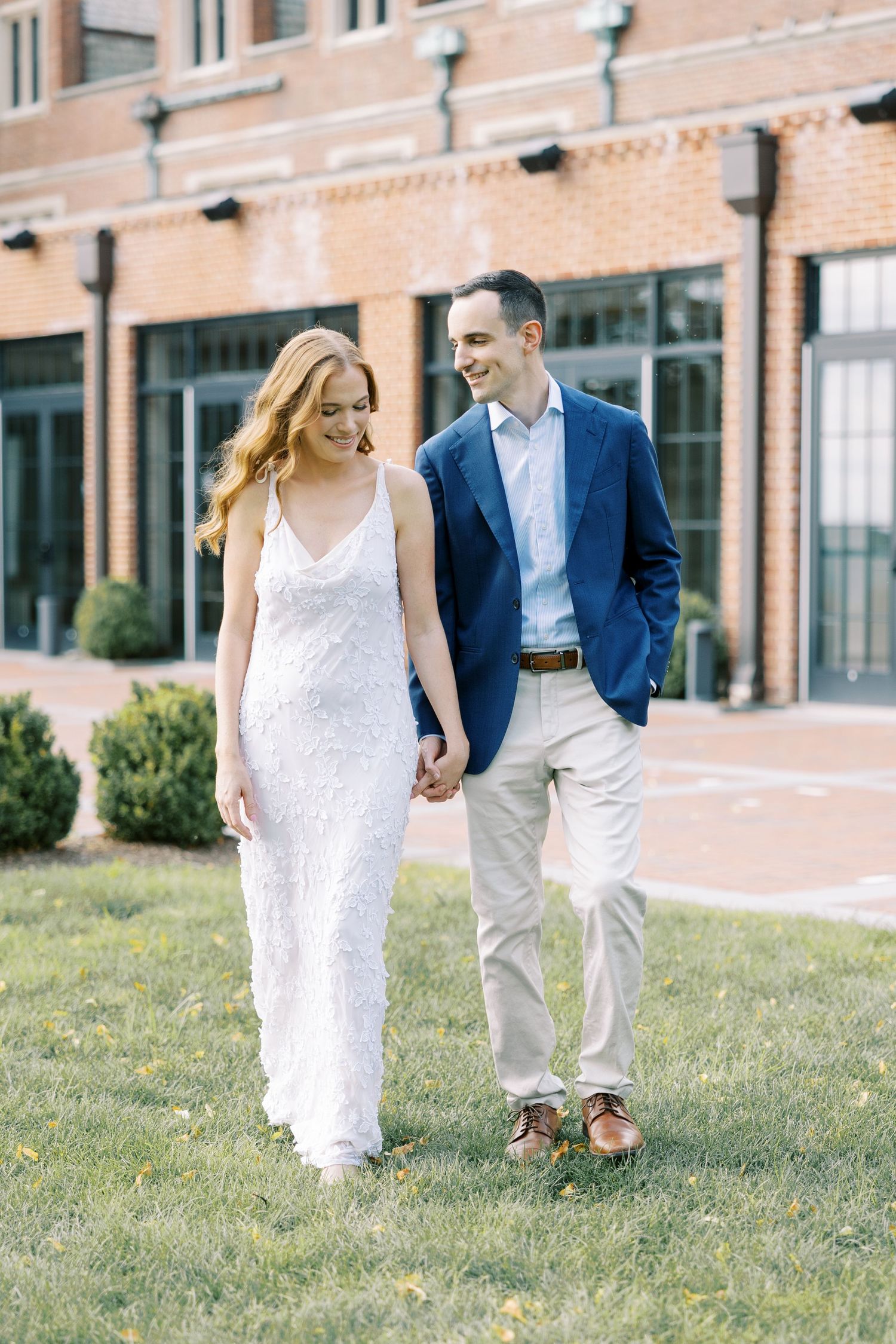 A couple walking on grass, with the person in a white dress and their partner wearing a navy blazer near a brick building at Pendry Natirar.